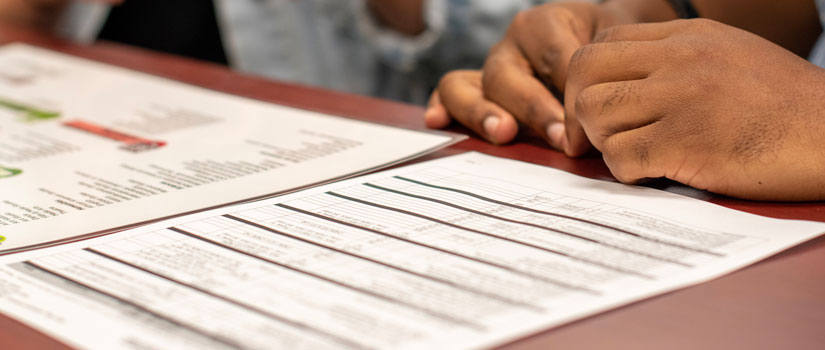 In the foreground a major map with a list of suggested courses is shown next to a list of UofSC majors. In the Background an advisors brown hands are clasped.