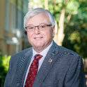 Dr. Amiridis is pictured from the shoulders up wearing a black and white jacket, garnet tie, and glasses. He has pale skin, white hair and is smiling.