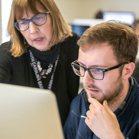 Male student working at computer with teacher looking over shoulder to offer help