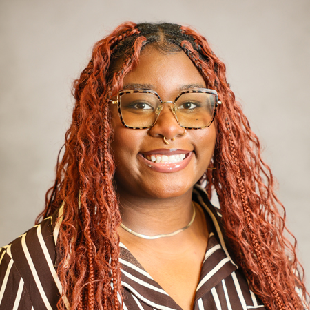 Professional headshot of a smiling person with long auburn braided hair wearing oversized tortoiseshell eyeglasses and a brown-and-white striped blouse. The person is facing the camera and is wearing a septum ring and a silver necklace. The photo is set against a soft gray studio background with warm, even lighting, creating a friendly, approachable, and professional appearance.