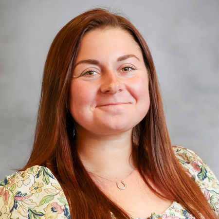 Portrait of a smiling woman with long auburn hair wearing a floral blouse against a gray background.