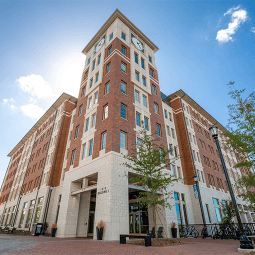 Students walking and on bikes walk on a brick courtyard. Behind them is a six story brick residence hall flanked by trees.