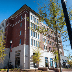 Students walking and on bikes walk on a brick courtyard. Behind them is a six story brick residence hall flanked by trees.