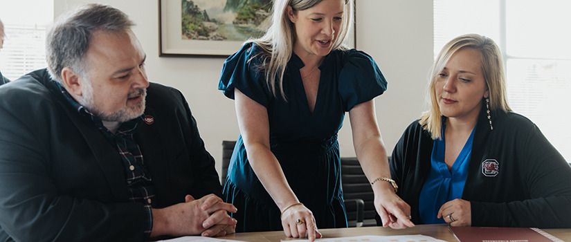 Male and two women are looking over papers on a table.