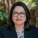  A professional headshot of a woman wearing glasses and a dark blazer, smiling slightly, with greenery softly blurred in the background.