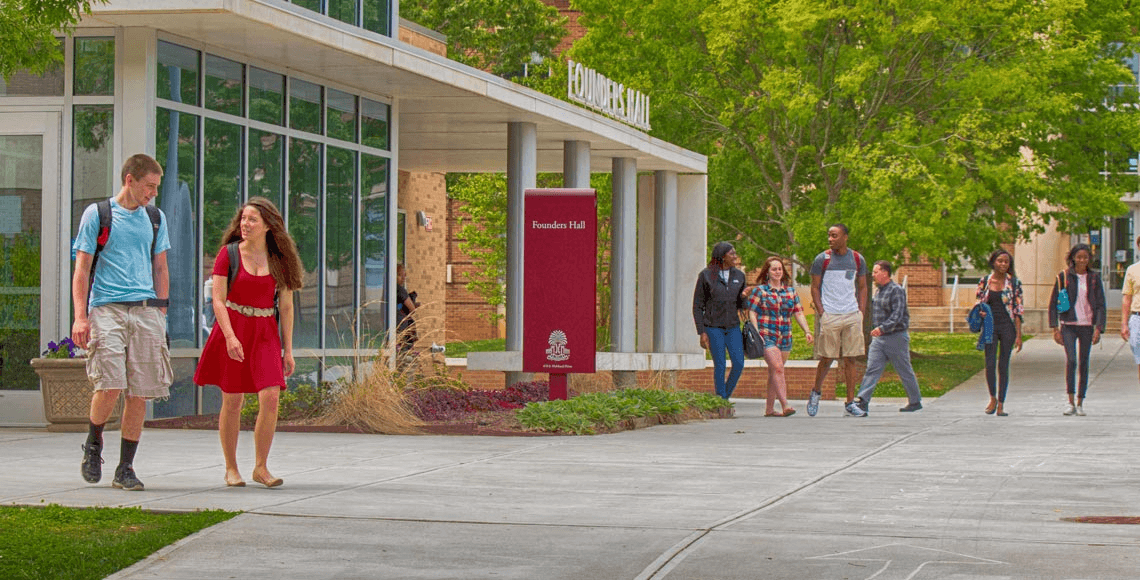 Groups of students wearing backpacks walk on the sidewalks in front of Founders Hall on the USC Lancaster campus