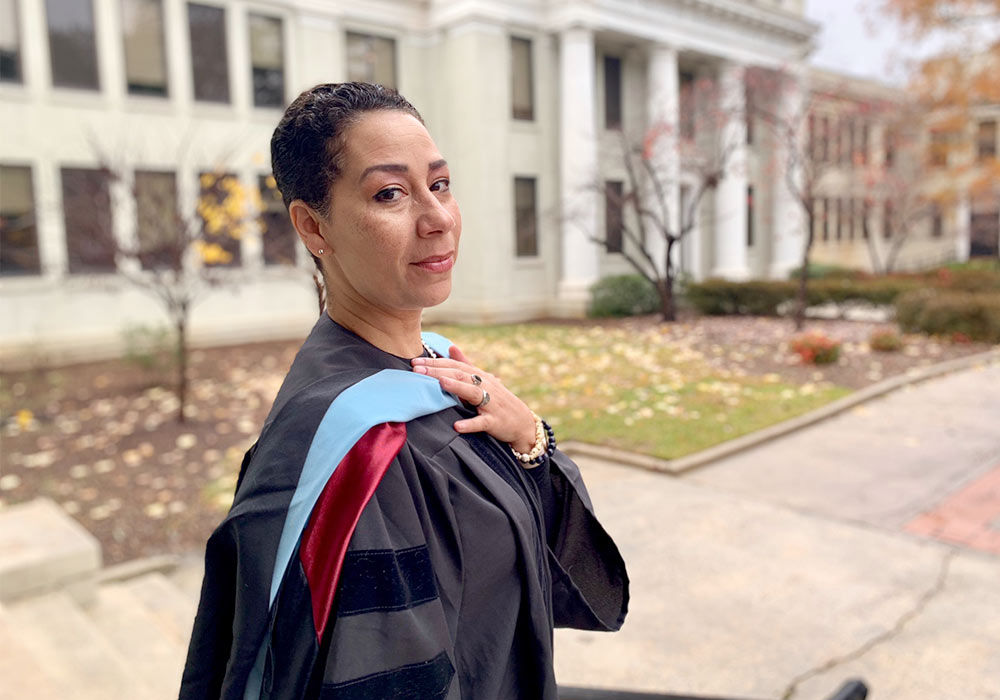 Wendy stands in the courtyard of the Education building looking over her shoulder with doctoral hood and graduation robes on.