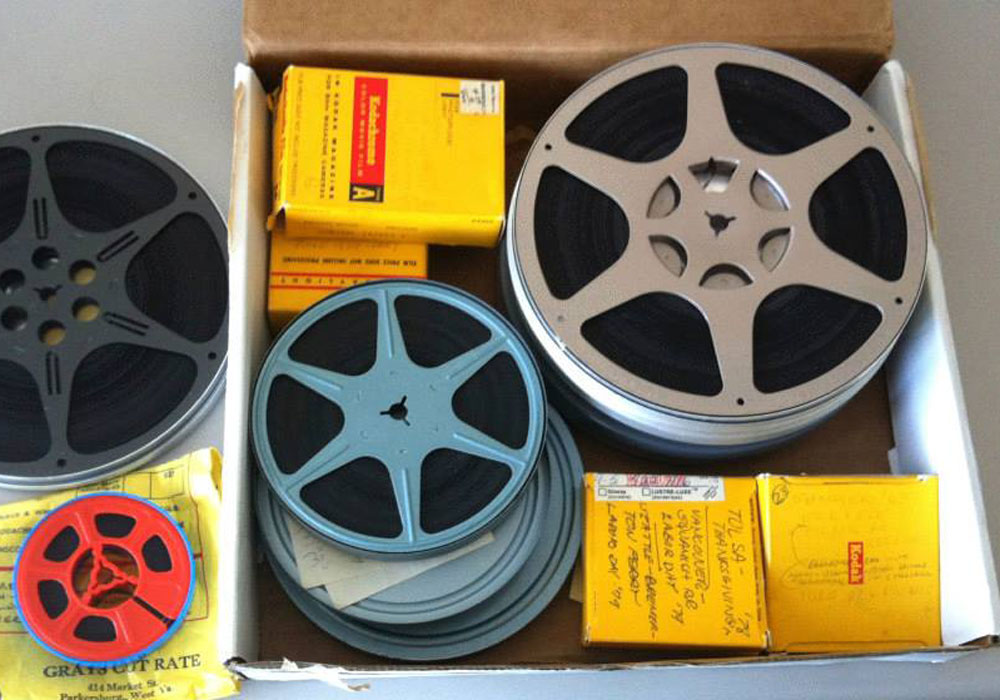 An open cardboard box filled with vintage film reels of various sizes and colors, alongside several yellow Kodak film boxes, viewed from above.