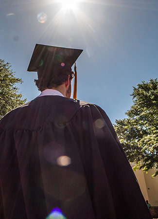 Student from behind wearing a graduation cap and gown with a blue sky in the background.
