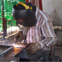 A figure wears a black welding helmet and holds welding equipment while sparks fly. Photo by John Doering-White.
