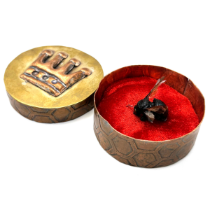 A preserved bee lays on a red velvet pillow in a handmade copper and brass container, etched with a honeycomb and crown motif