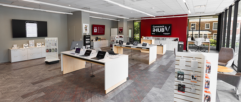 Interior of the Gamecock iHub featuring a central table with laptops on display, a red accent wall with the Gamecock iHub logo, and large windows overlooking an outdoor seating area.