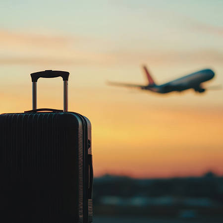 Black suitcase in the foreground with a commercial airplane taking off at sunset in the background.