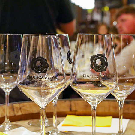A collection of wine glasses with the text Enoteca Alessi Delicatessen Dal 1952, Firenze in the foreground and patrons sitting at a table in a blurred background.