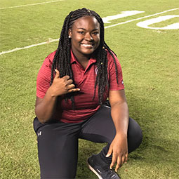 Lyric Swinton kneels on a football field and gives the spurs up hand signal