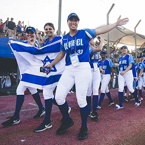 Israel softball players celebrate on the field, holding a national flag during an international competition.
